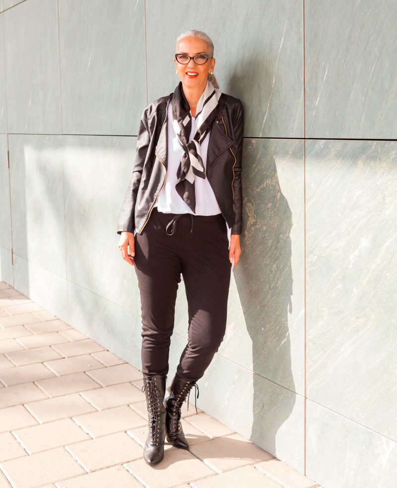 Dojrzała kobieta w stylizacji smart casual: czarna ramoneska, materiałowe spodnie, kowbojki z biała koszulą i czarno białą apaszką