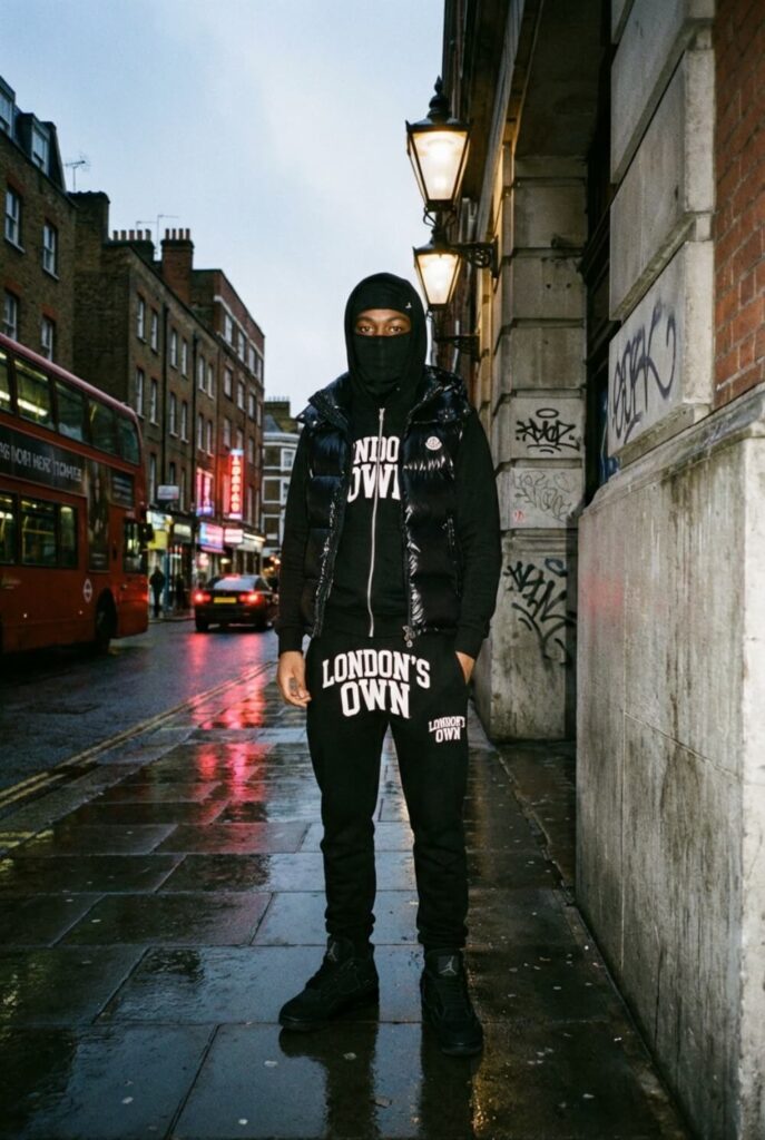 Uliczne ujęcie mężczyzny w czarnym dresie z pikowaną kamizelką, kominiarką i masywnymi butami w stylu Jordan