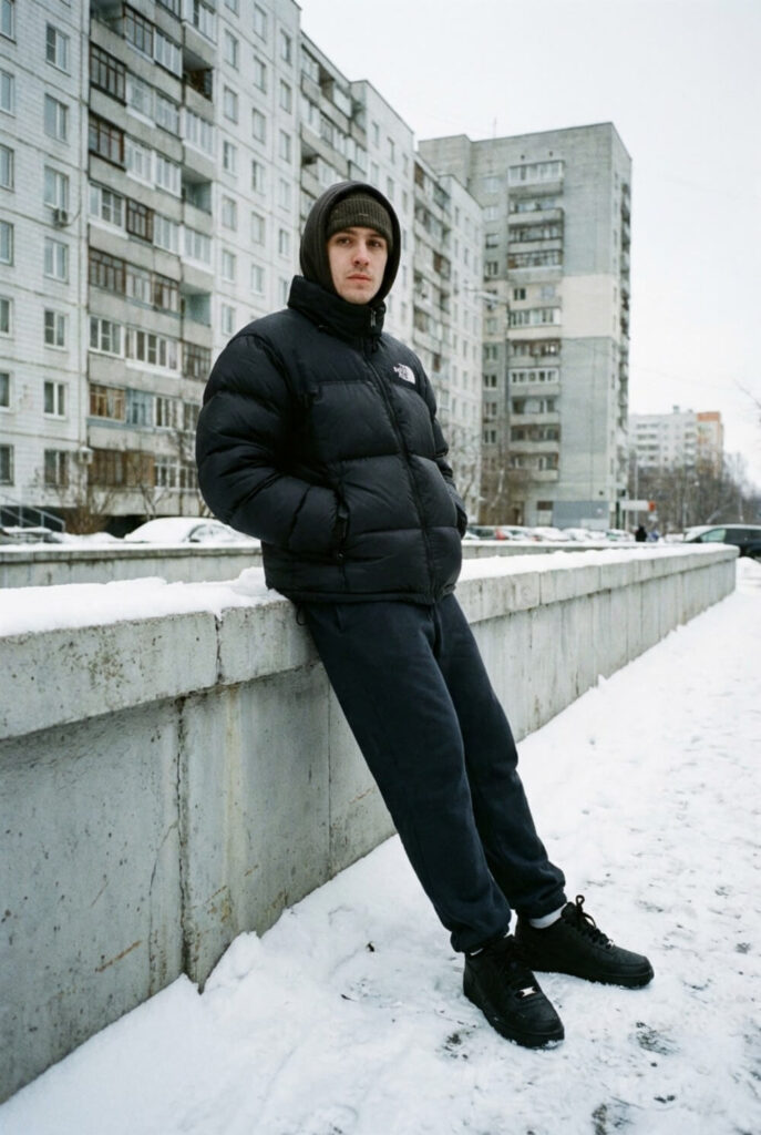 Zimowe ujęcie mężczyzny w napompowanej kurtce puchowej, ciemnym dresie i czarnych sneakersach na tle bloków