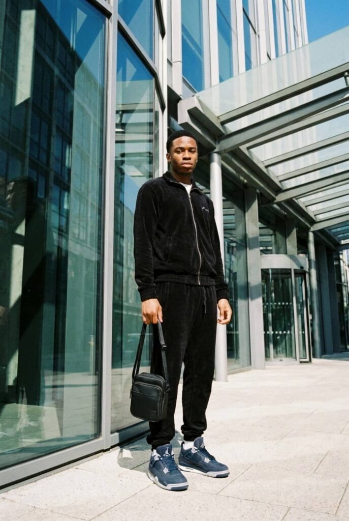 Mężczyzna w czarnym dresie i butach w stylu Air Jordan 4, trzymający dopasowaną torbę na tle nowoczesnej architektury