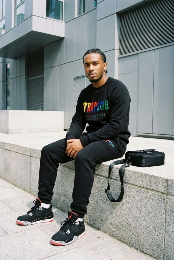 Półzbliżenie mężczyzny w czarnym dresie hypebeast z butami w stylu Jordan 4 i dopasowaną torbą na tle nowoczesnych budynków