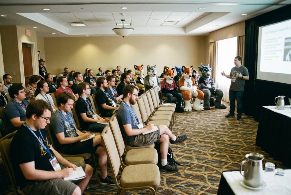 Prelekcja na konwencie furry w hotelowej sali konferencyjnej, z uczestnikami i kilkoma fursuiterami
